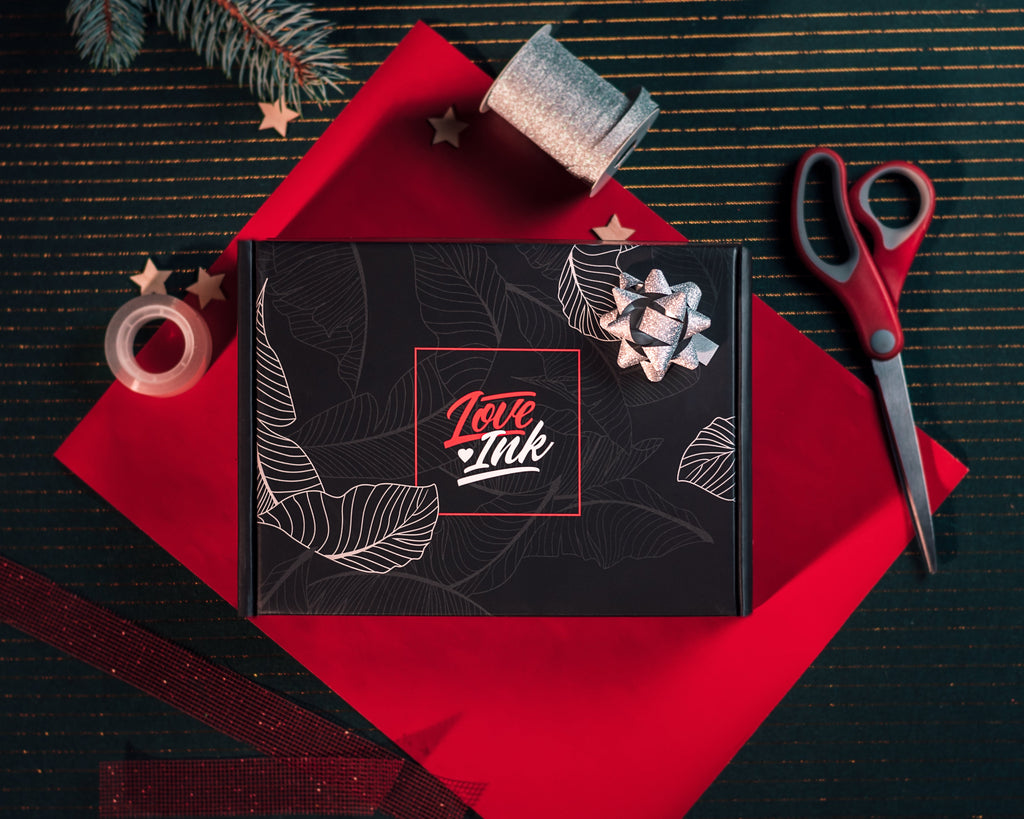 Černá dárková krabička s logem Loveink leží na červeném balicím papíře vedle nůžek, role stříbrné stuhy a červené dekorační pásky na tmavém podkladu s jehličím a vánočními hvězdami.