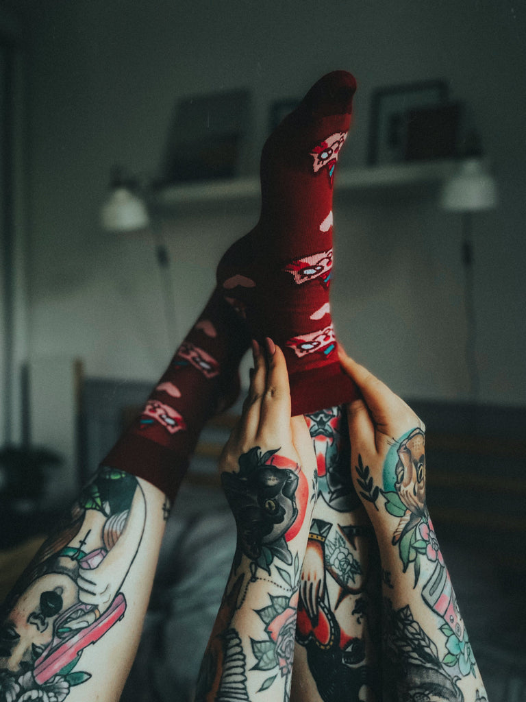 Osoba w vínových ponožkách s motivem kočorožce, sedící s nohama vztyčenými nahoru, v pokoji s tetováním zdobícím nohy i ruce.