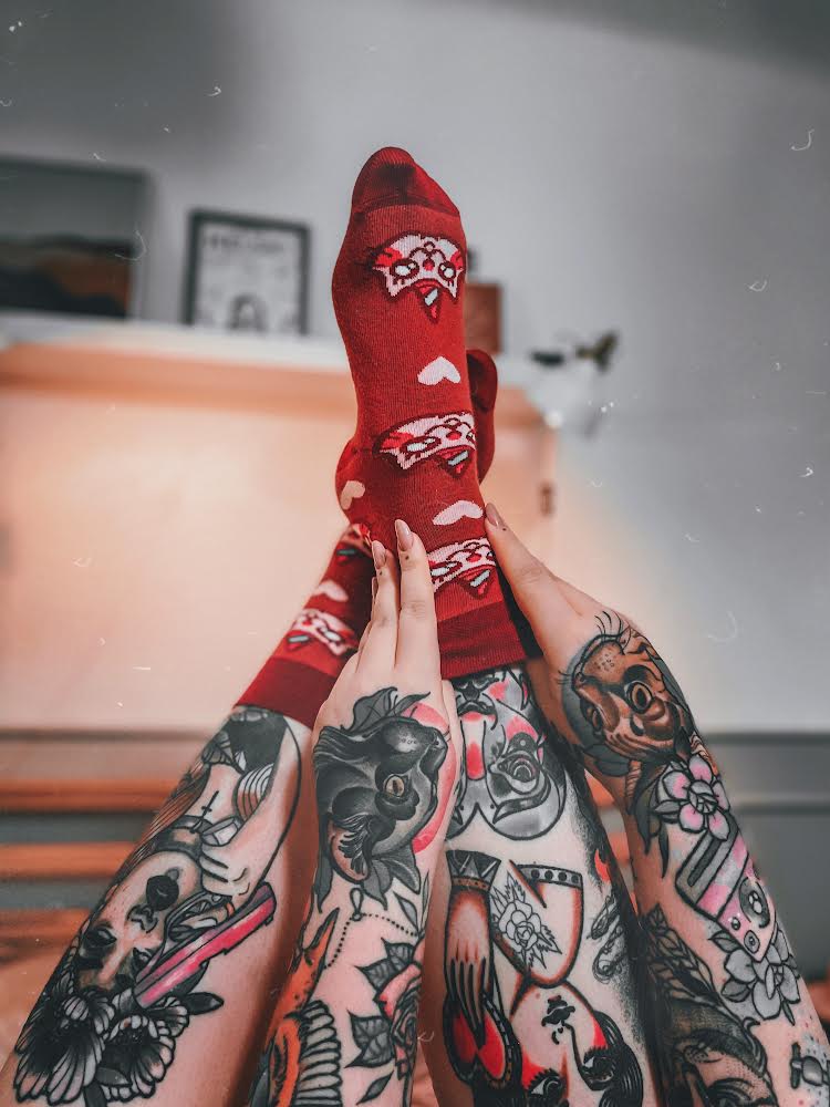 Osoba w vínových ponožkách s motivem kočorožce, sedící s nohama vztyčenými nahoru, v pokoji s tetováním zdobícím nohy i ruce.