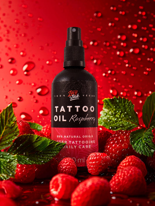 Na obrázku je láhev tetovacího oleje s malinovou vůní od značky Love Ink. Láhev je černá s růžovou etiketou a nápisem "Tattoo Oil Raspberry". Kolem láhve jsou čerstvé maliny a zelené listy na červeném pozadí s kapkami vody, což dodává celkovému vzhledu svěžest a přirozenost. Olej je označen jako 100% přírodní a veganský, určený pro každodenní péči po tetování.