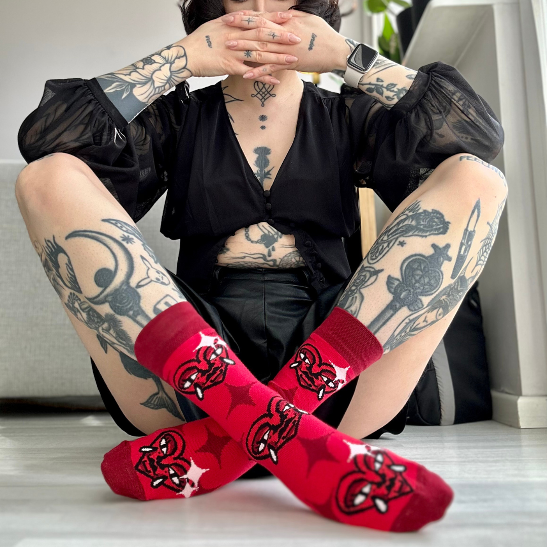 Osoba s tetováními sedí s nohami zkříženými a ukazuje červené ponožky Loveink s motivem "heart", vytvořeným designérem Fat Kat.