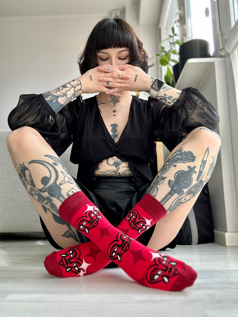 Osoba s tetováními sedí s nohami zkříženými a ukazuje červené ponožky Loveink s motivem "heart", vytvořeným designérem Fat Kat.