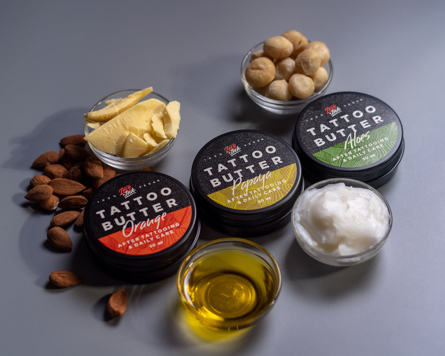Tři kelímky s Tattoo Butter různých druhů - 'Orange', 'Papaya' a 'After Inked', rozmístěné vedle skleničky se žlutým olejem, hromádky mandlí a skupinky lískových ořechů na šedém pozadí.