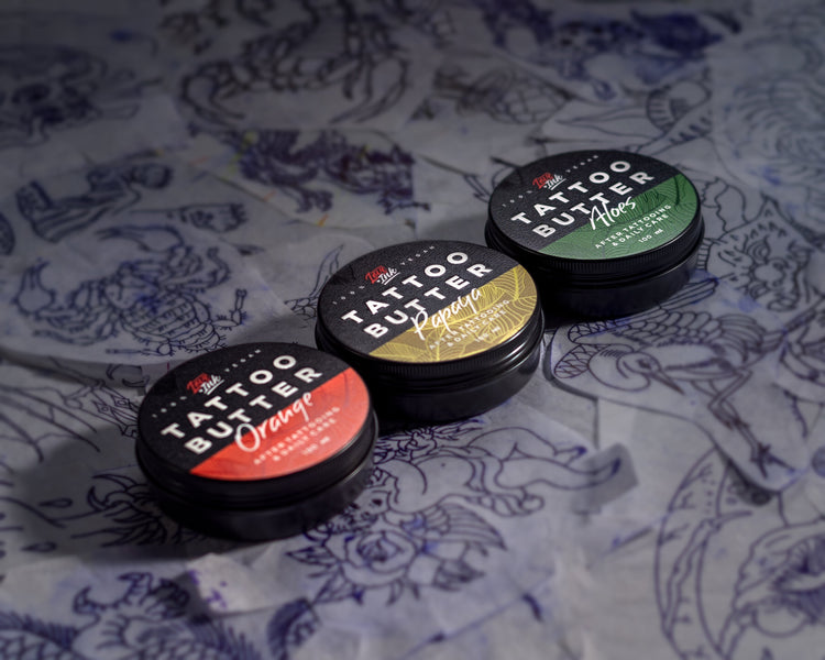 ři 100ml kelímky s Tattoo Butter různých druhů - 'Orange', 'Papaya' a 'Aloes', rozmístěné vedle skleničky se žlutým olejem, hromádky mandlí a skupinky lískových ořechů na šedém pozadí.