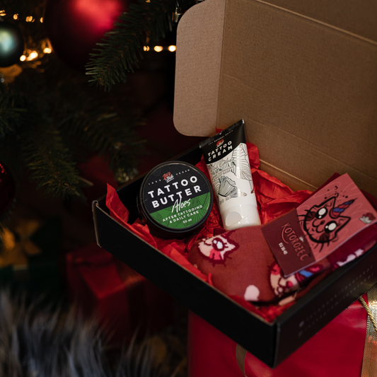 set Love Ink v černé krabičce, obsahující zelenou plechovku Tattoo Butter Aloes, bílý tubu s Tattoo Cream Pina Colada a páry červeno-bílých ponožek s obrázky kočiček, prezentované na červeném hedvábném papíře