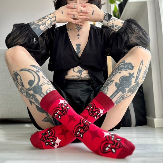 Osoba s tetováními sedí s nohami zkříženými a ukazuje červené ponožky Loveink s motivem "heart", vytvořeným designérem Fat Kat.